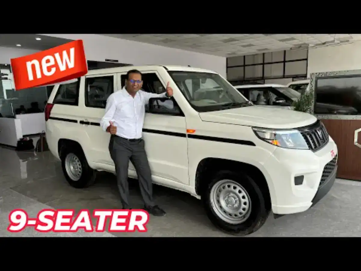 White 9-seater SUV with a man in business attire posing next to it, inside a showroom. Bold "new" text on the top left corner.